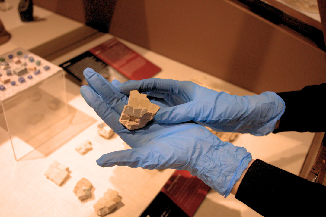 Gloved hand holding fragment of a mosaic over an exhibit case